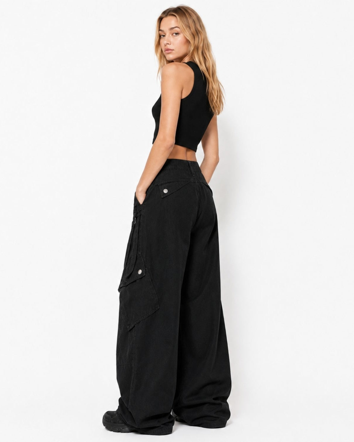 A woman stands in profile wearing a black sleeveless crop top and very wide-leg black cargo pants against a clean white studio background.