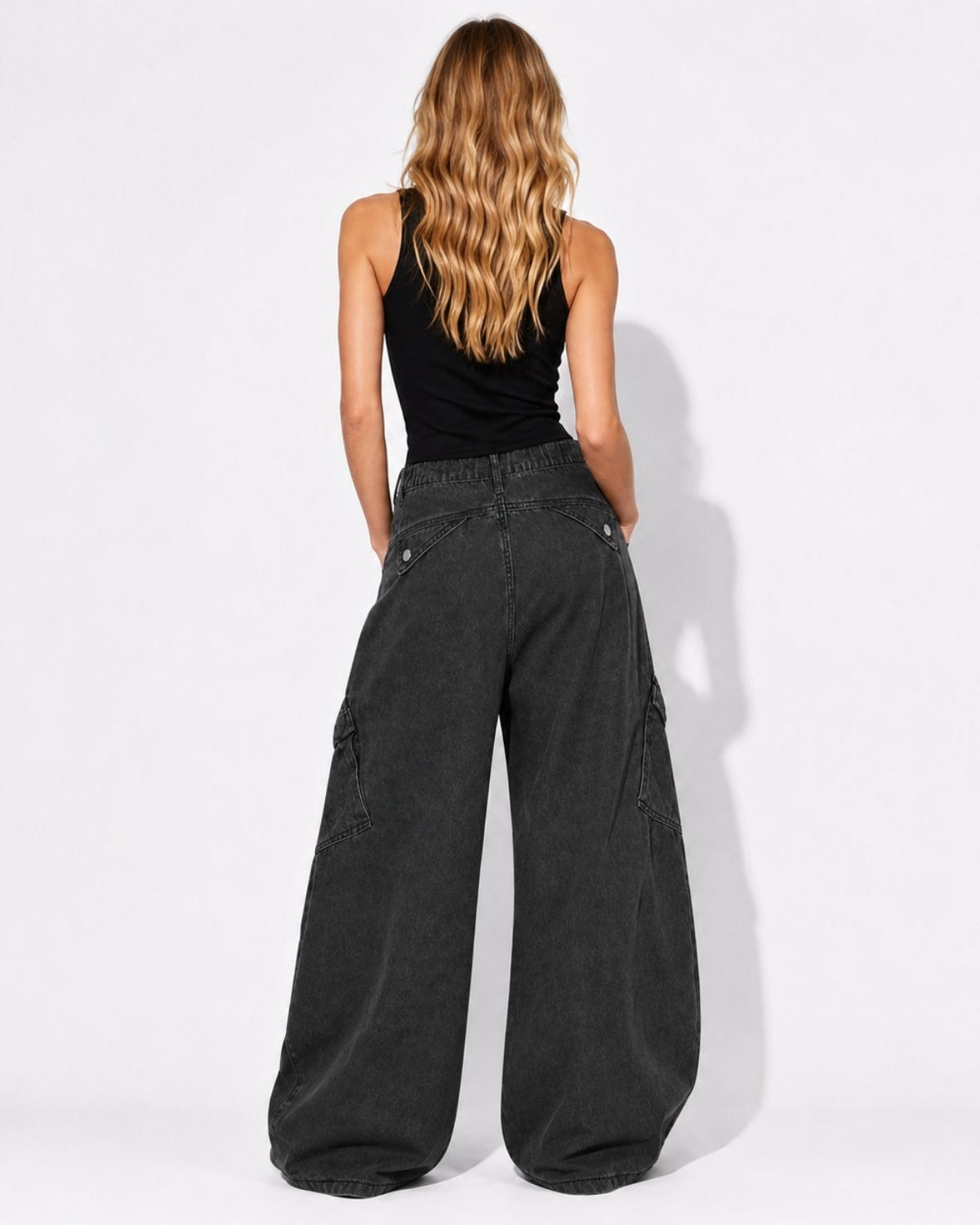 A woman viewed from behind wearing wide-leg charcoal gray cargo pants and a black tank top, standing against a plain white studio background.