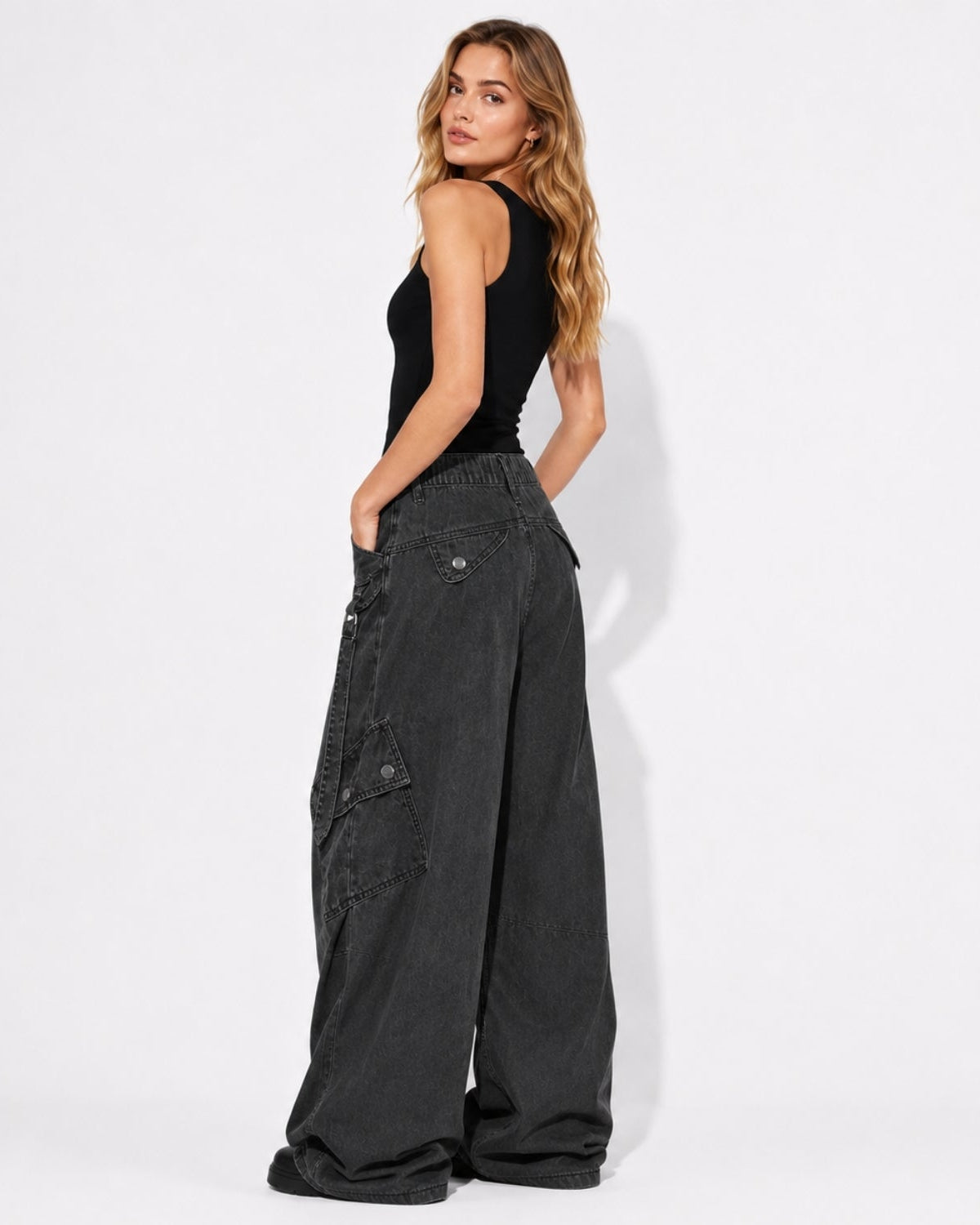 A woman stands in profile wearing a black sleeveless top and very wide-leg dark charcoal cargo pants against a clean white studio background.