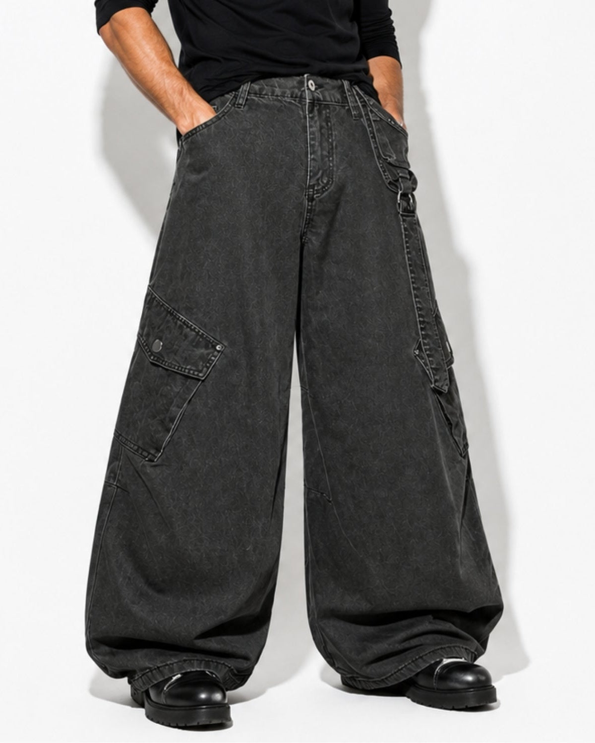 A person wearing oversized dark charcoal cargo pants with side flap pockets, a black long-sleeve top, and black shoes, standing against a plain light background.