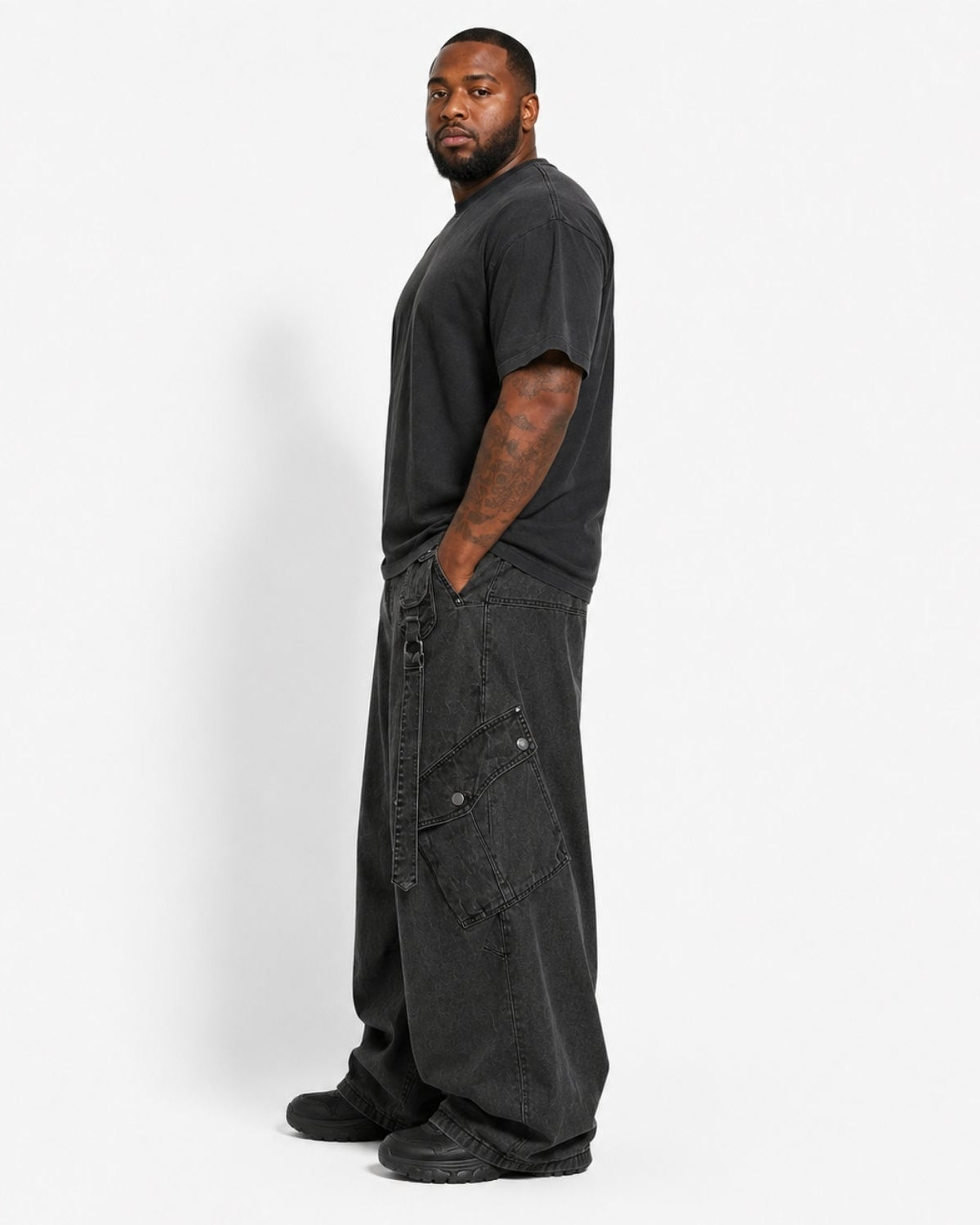 A man wearing a black crewneck t-shirt and oversized black cargo pants, standing in profile against a light gray studio background.