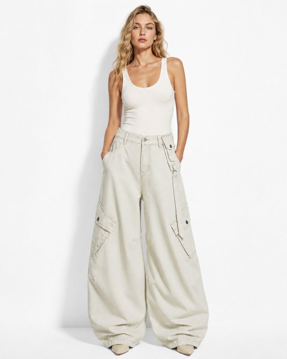 A woman modeling a pair of oversized, wide-leg cargo pants in a light beige color, paired with a white ribbed tank top, standing against a plain white studio background.