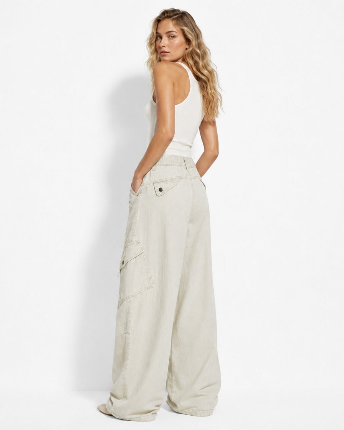 A woman stands in profile wearing a white ribbed tank top and wide-leg cargo pants in a light oatmeal beige color, photographed against a clean white studio background.