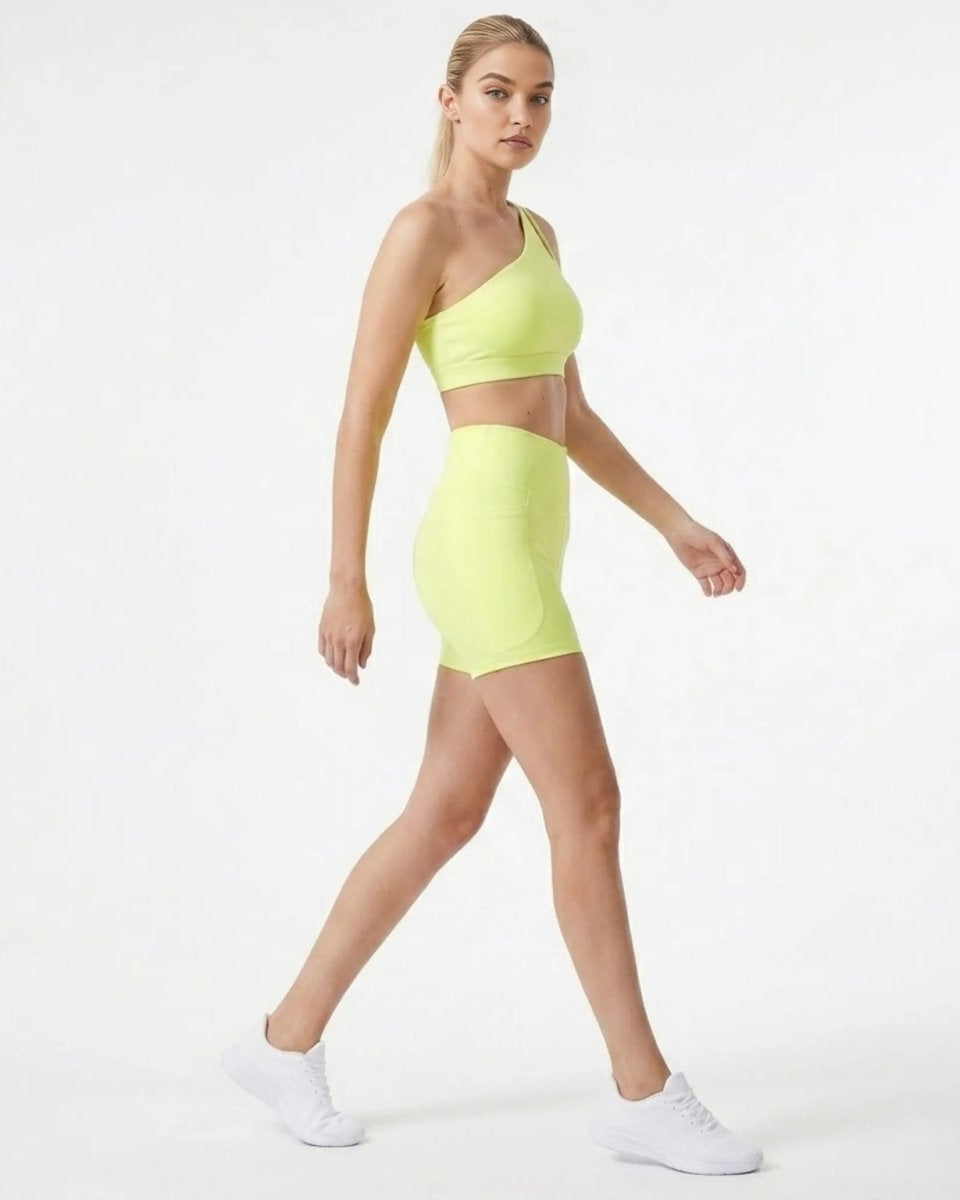 Side profile of model walking in premium yellow matching workout set featuring high rise pocket shorts and one shoulder crop top