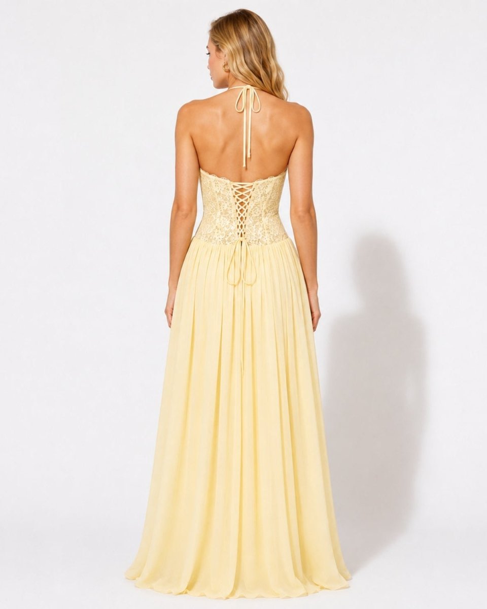 A woman shown from the back wearing a pale yellow halter maxi dress with a lace-up open back and a flowing chiffon skirt, photographed against a plain white studio background.