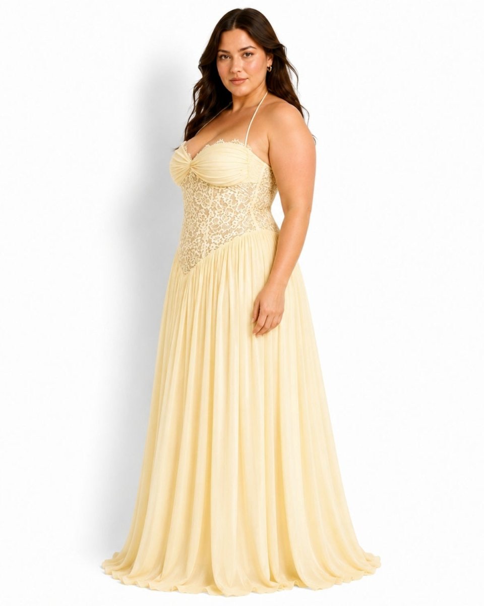 A woman modeling a floor-length pale yellow formal gown with thin straps, a ruched sweetheart bodice, and a lace-embellished waistband against a plain white studio background.