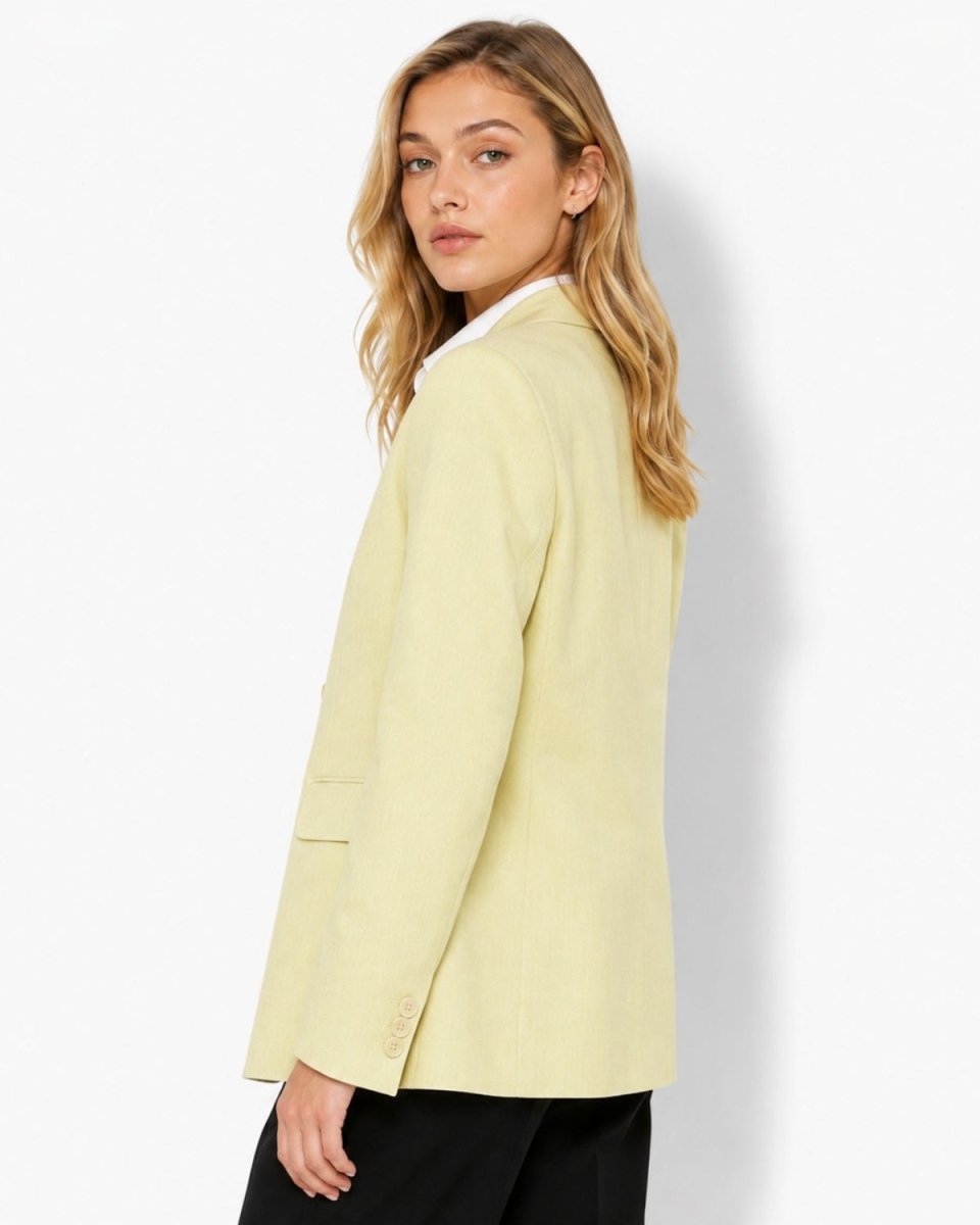 A woman wearing a pale yellow blazer over a white collared shirt and black pants, photographed from the side against a clean white studio background.