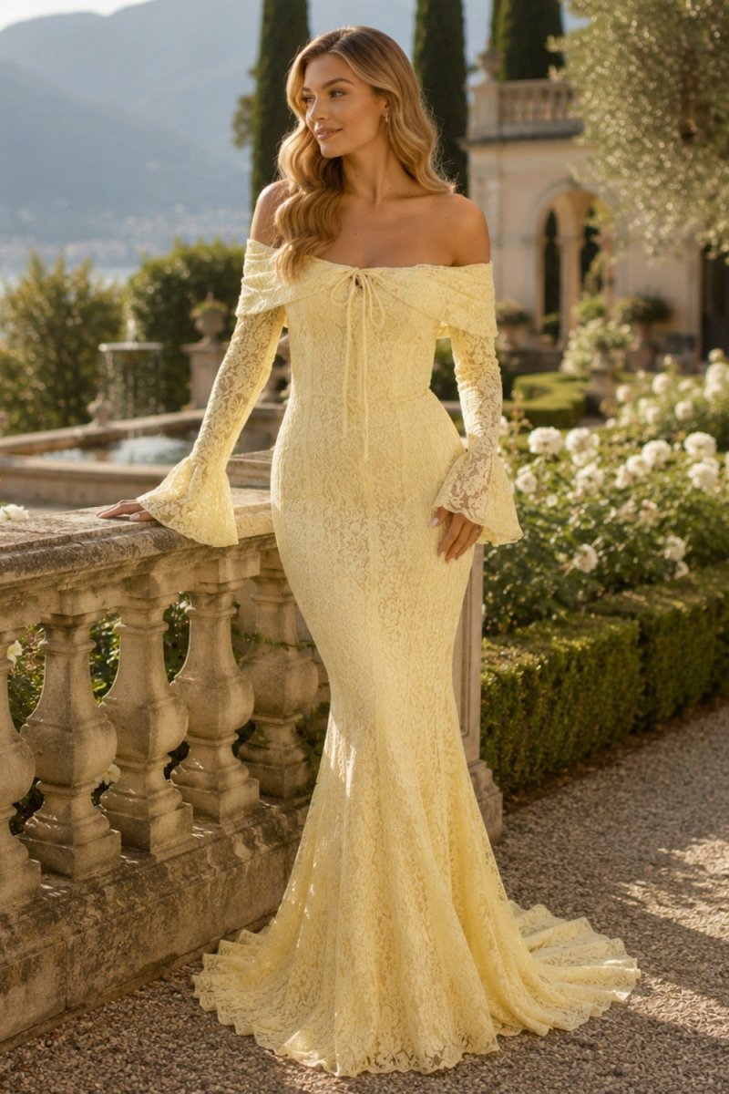 A woman wearing a pale yellow off-the-shoulder lace mermaid gown with long bell sleeves, standing outdoors in a garden setting next to a stone balustrade.