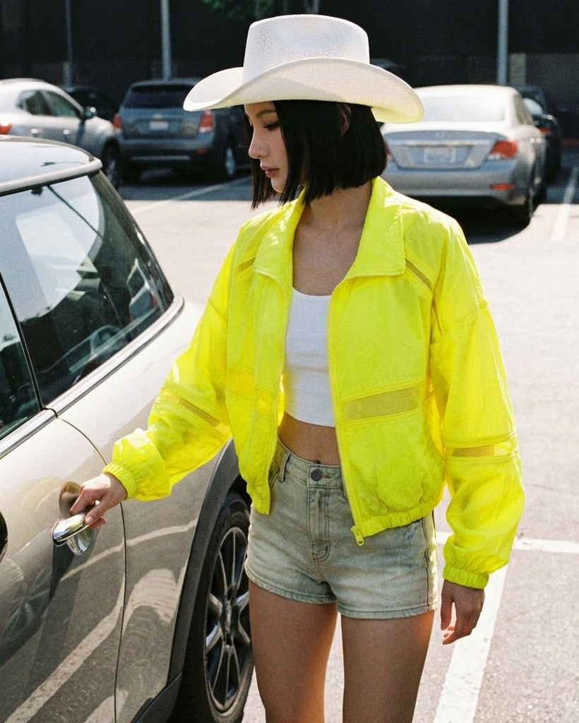  Lifestyle shot of woman opening car door wearing neon yellow mesh panel windbreaker layered over a white crop top and denim shorts.